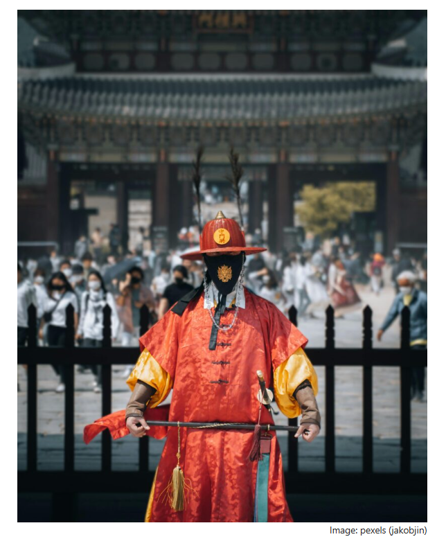 A royal guard in traditional red Joseon-era uniform standing in front of a palace gate, with visitors blurred in the background.