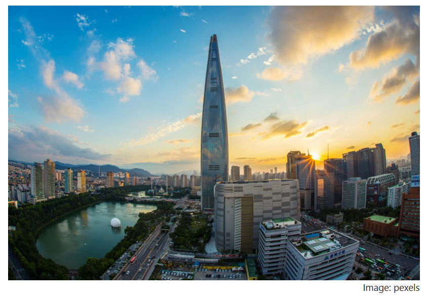 Sunset view of Seoul with a tall glass skyscraper rising above the city, surrounded by dense buildings, a river, and evening light.