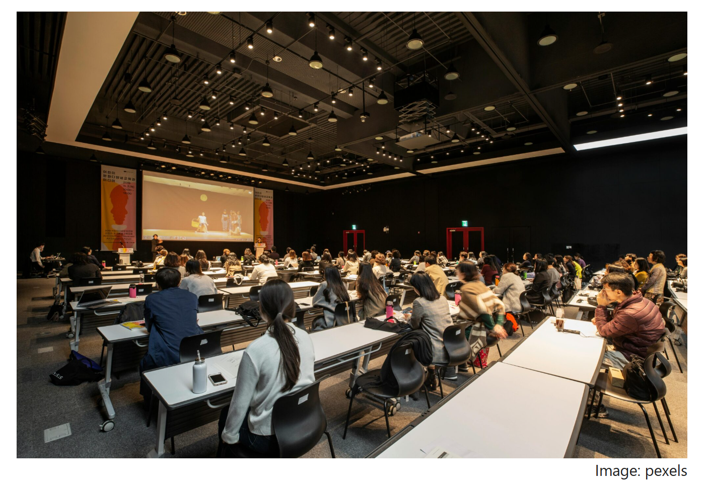 A large lecture hall with many people seated at desks, watching a presentation projected on a screen at the front.